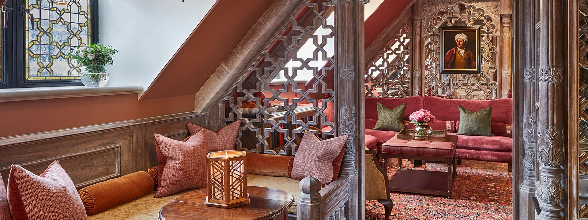 The King's Lodge: a cosy, elegant seating area with a warm colour palette, featuring cushioned benches, intricate lattice-like wooden partitions, and a framed portrait hanging over a red sofa.