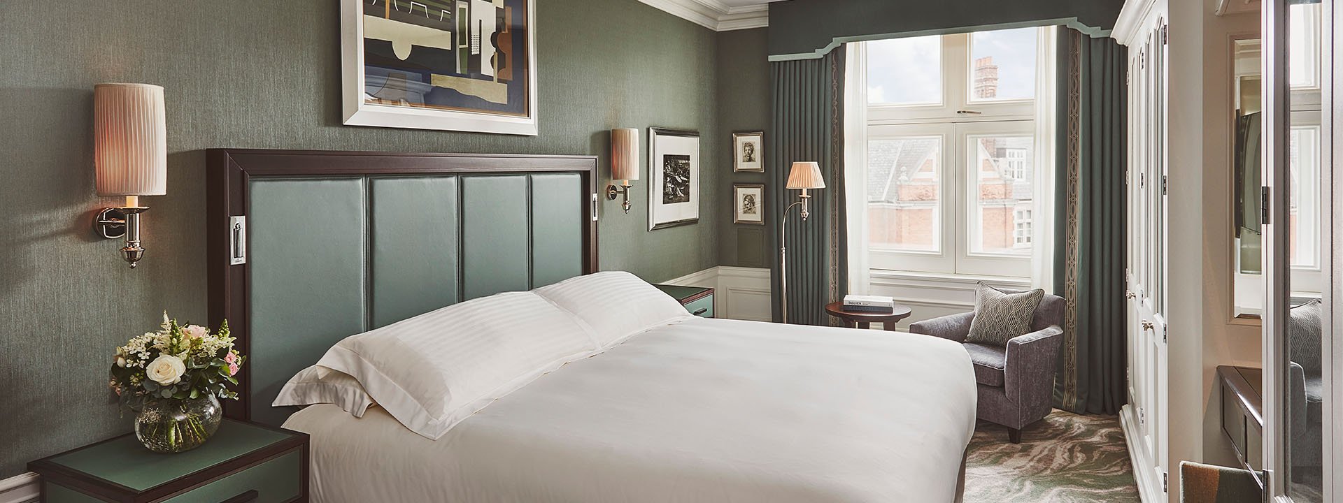 Coburg Suite: modern, minimalist hotel bedroom with muted green walls, a large bed with white linens, and a comfortable sitting area by the window.